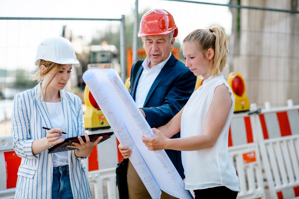Zwei Ingenieurinnen und ein Ingenieur prüfen Baupläne auf einer Baustelle in Köln mit Sicherheitshelmen.