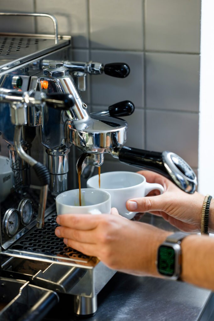 Zwei Kaffeetassen werden von einer professionellen Espressomaschine gefüllt, um die urbane Caféästhetik und den Genuss von Köln zu unterstreichen.