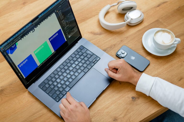 Nahaufnahme eines Laptops mit geöffnetem Design-Programm auf einem Holztisch; daneben Smartphone und Kopfhörer als Zeichen für modernes Arbeitsumfeld.
