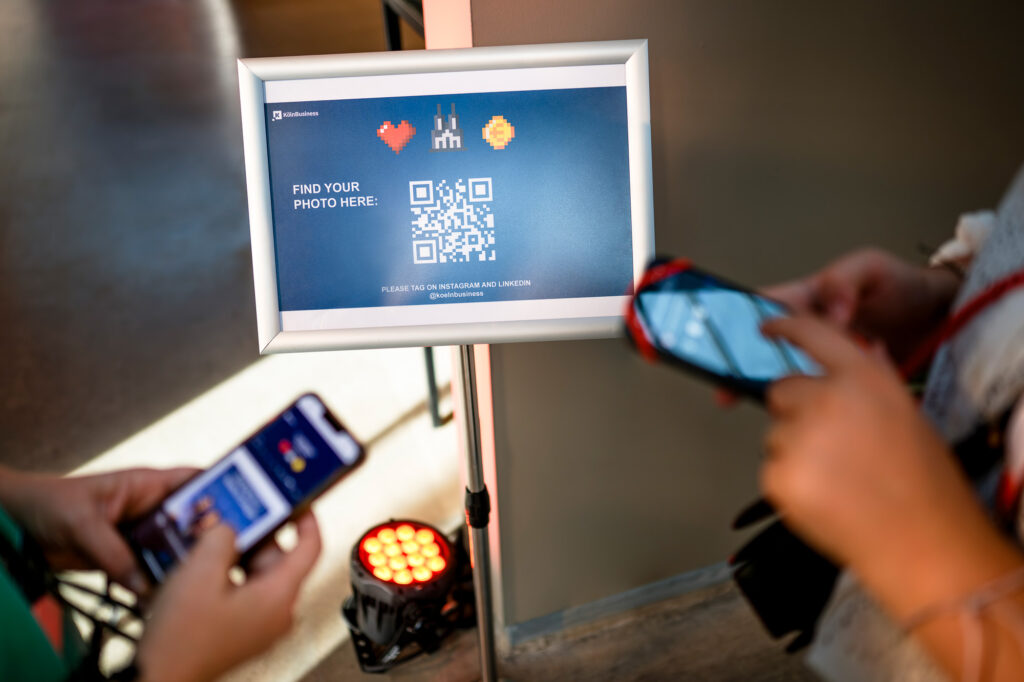 Zwei Personen scannen einen QR-Code auf einem Display bei einem Event in Köln.