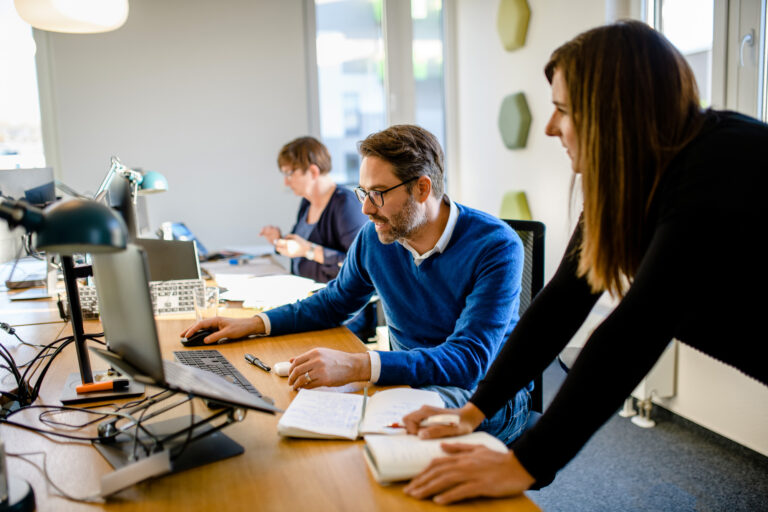 Drei Personen arbeiten zusammen in einem modernen Büro, mit Fokus auf einen Mann im Gespräch mit einer Kollegin am Tisch.