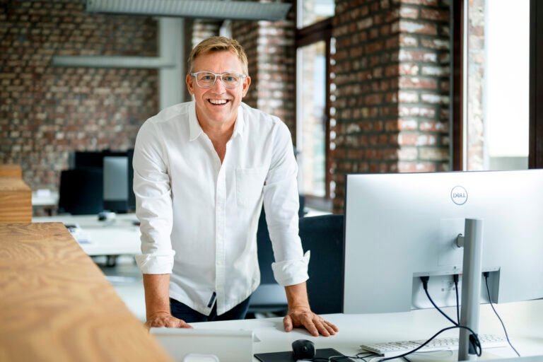Lächelnder Mann in modernem Büro mit Computern, repräsentiert professionelle Businessfotografie für Unternehmen.