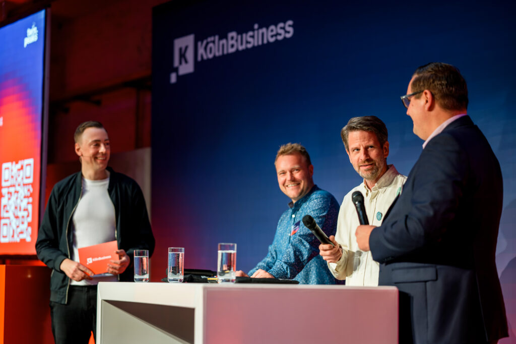 Drei Männer auf einer Podiumsdiskussion bei KölnBusiness, diskutieren Unternehmensstrategien und Innovationen im urbanen Umfeld.